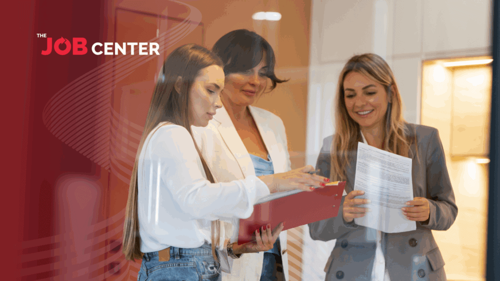 Woman in corporate attire holding a printed form discusses resumes with coworkers