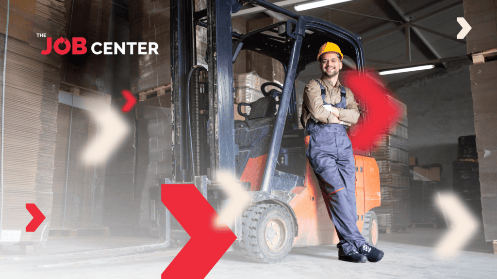 Man in safety hat and overalls stands smiling next to a forklift